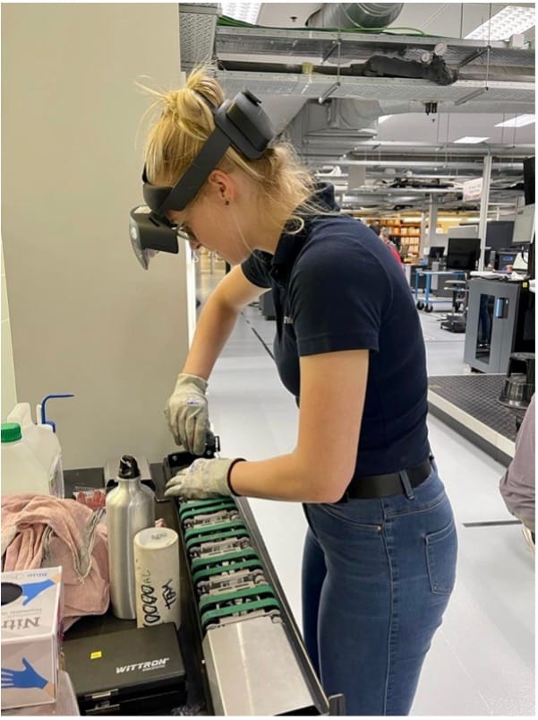 Vrouw werkt aan een transportband in een technische productieomgeving met veiligheidsbril en handschoenen